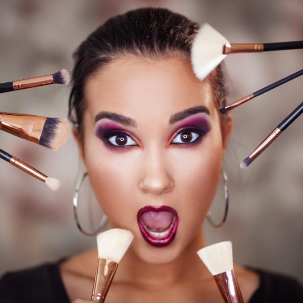 A portrait of a funny young woman with beautiful eyes, who is holding makeup brushes and representing a nice makeup on her face.