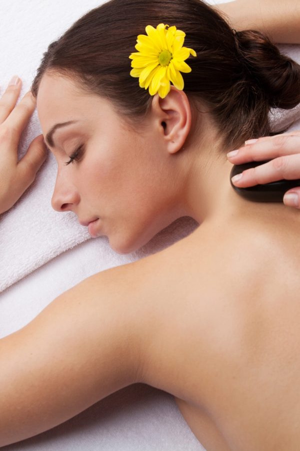 Stone treatment. Top view of attractive young woman with flower in head lying on front and keeping eyes closed while massage therapist massaging her back with spa stones